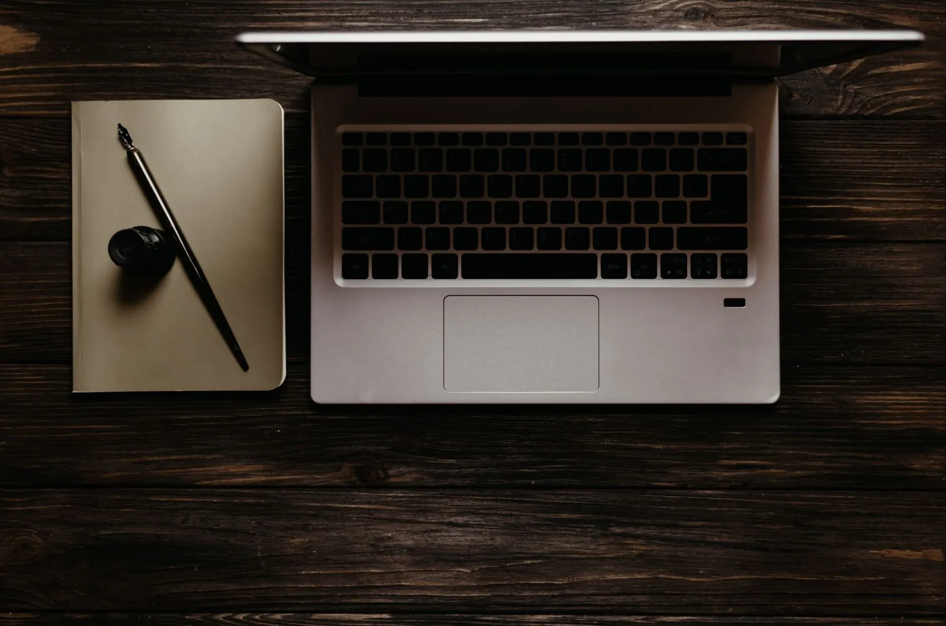 An open laptop and a notebook with a dip pen on a dark wooden desk, seen from above